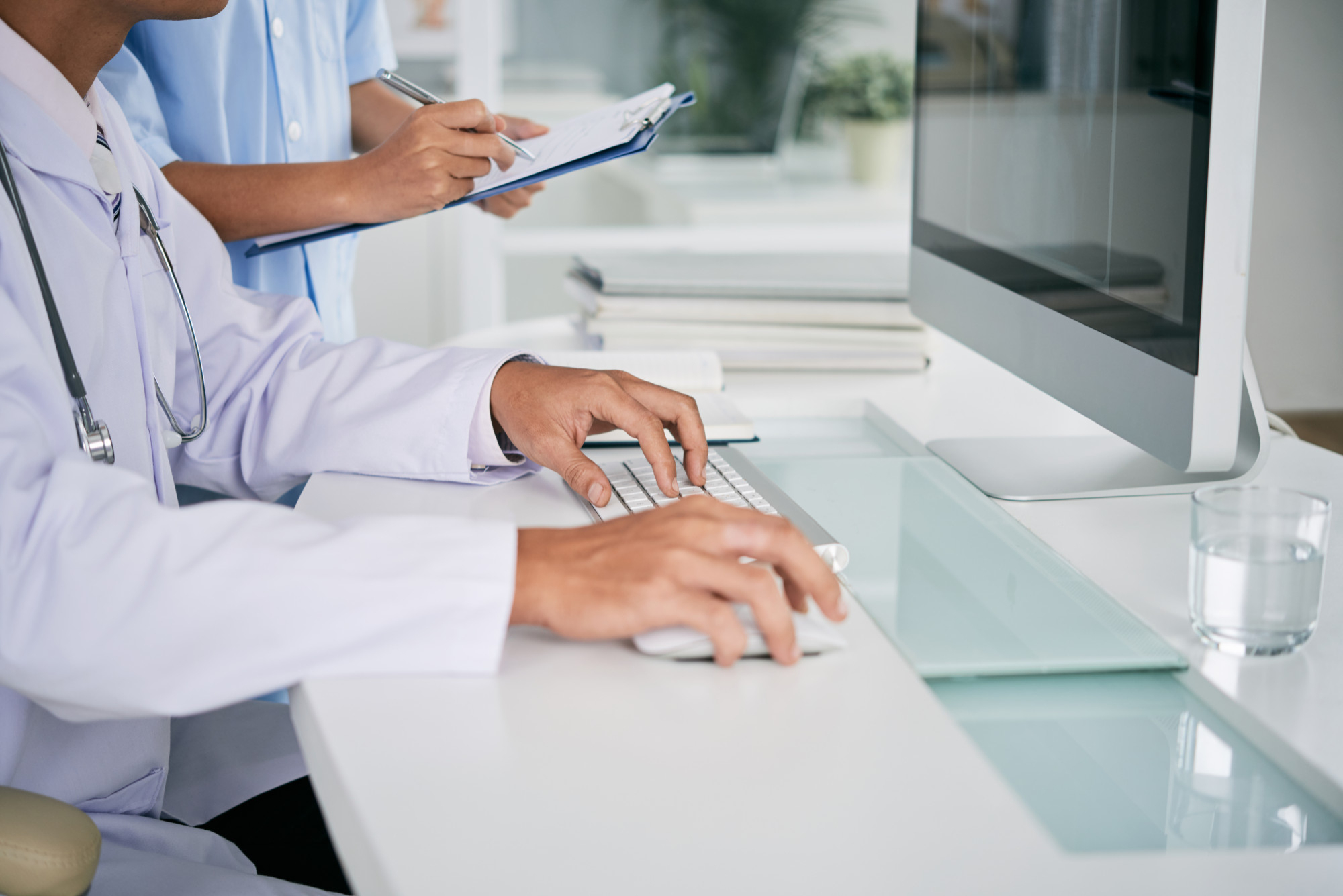 Doctor typing on a computer while another takes notes in a medical office.