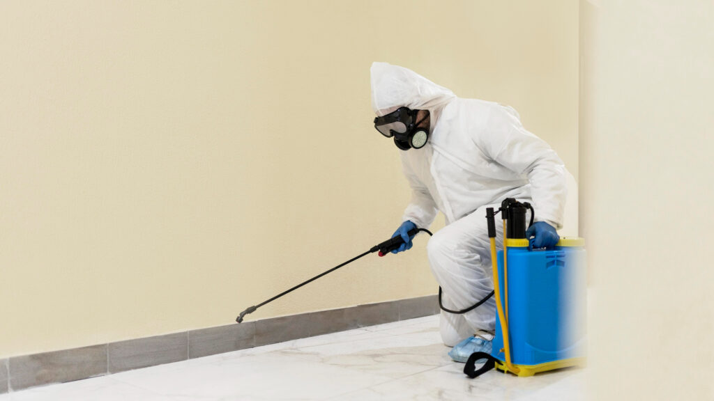 Worker in hazmat gear spraying pesticide along the base of an indoor wall.