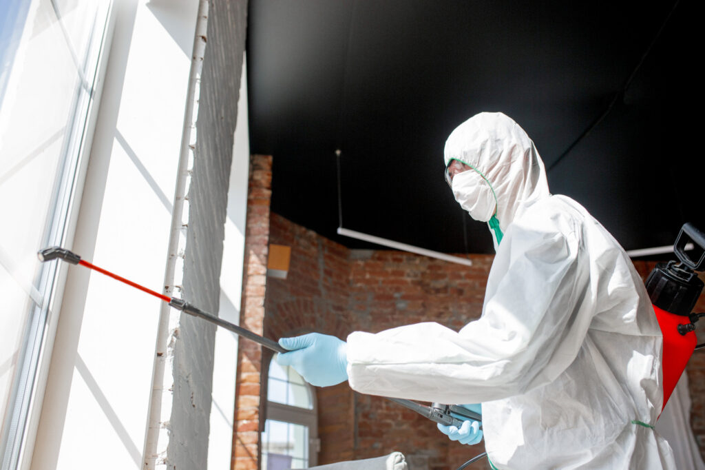 Technician in hazmat suit spraying disinfectant on a brick wall indoors.