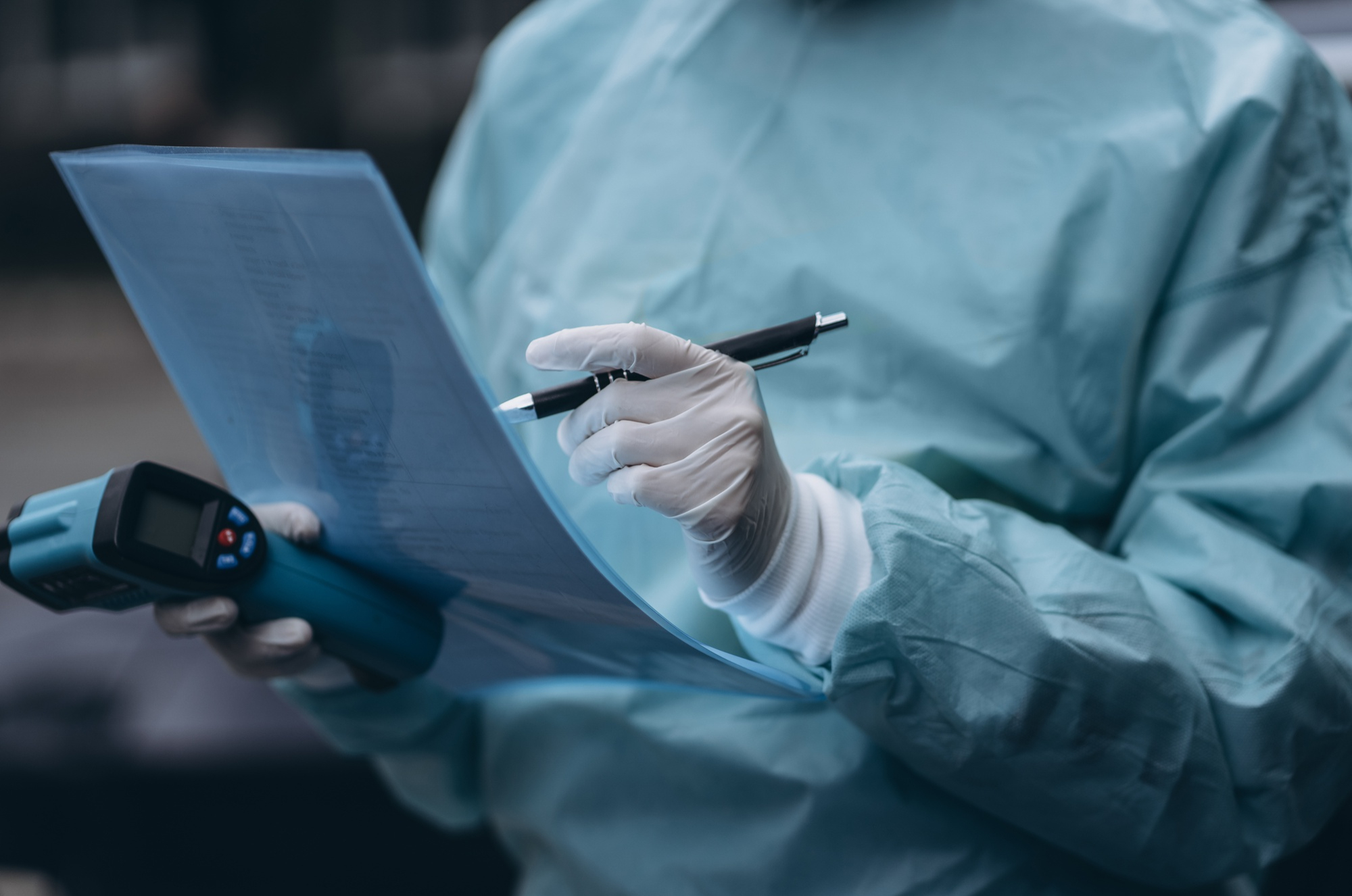 Healthcare worker in PPE writing notes with an infrared thermometer.